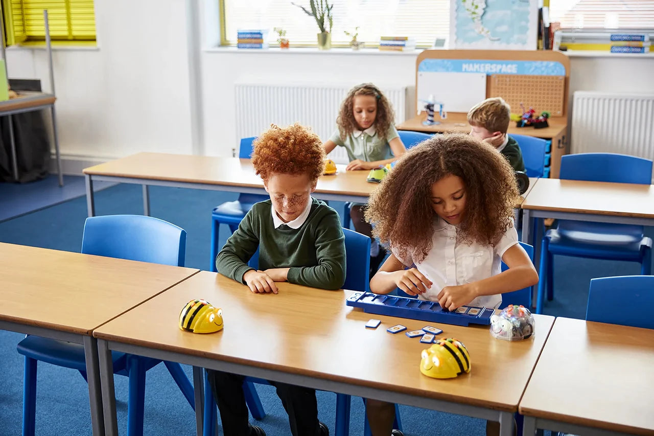 Children playing with toy robots at school desks in a bright classroom. TTS Products.
